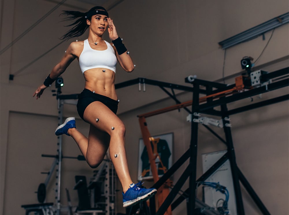 A female athlete, with 3D motion capture markers on her body, is sprinting in a gym.